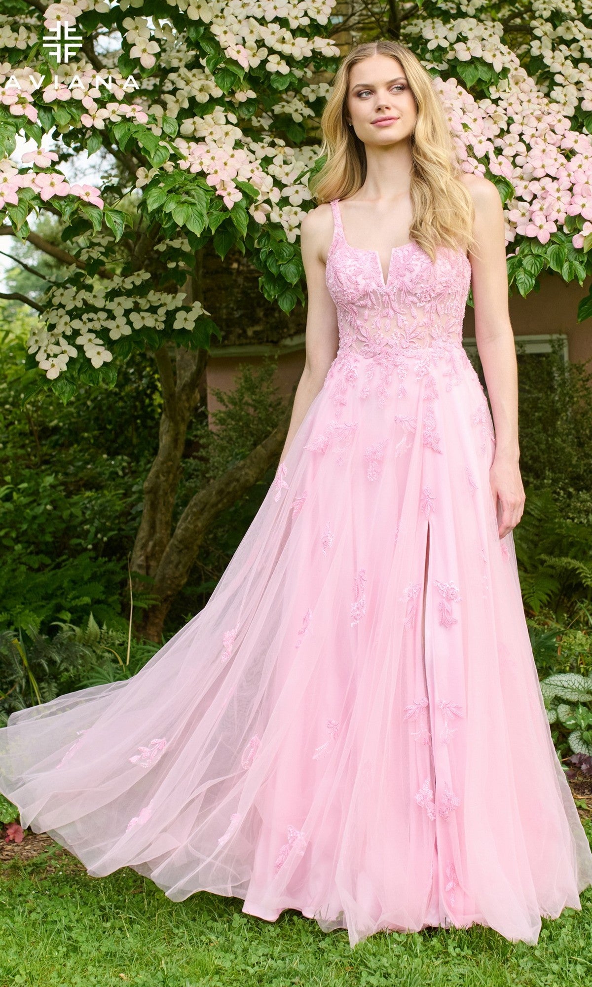 A woman stands outdoors before a flowering tree, wearing the Faviana 11437 long prom dress in light pink tulle with delicate lace applique.